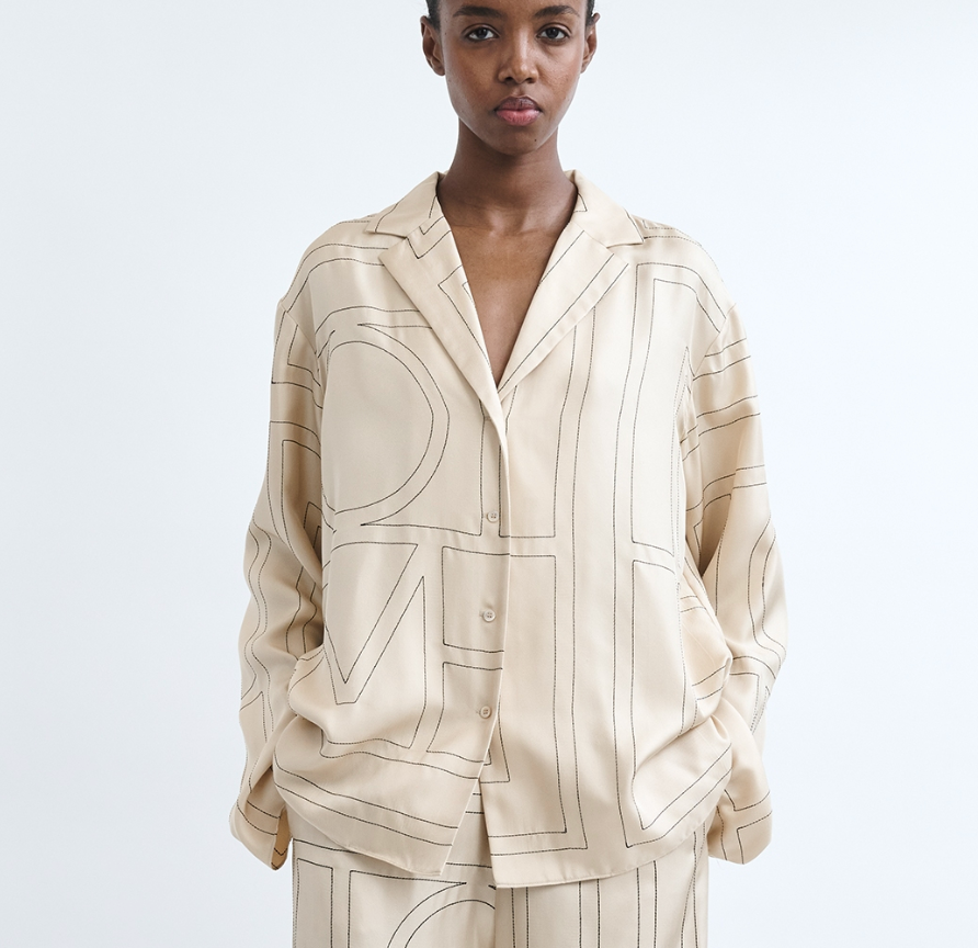 A person wearing the Toteme Monogram Silk PJ Top, a loose-fitting beige piece with a bold large-letter monogram pattern, stands against a plain white background.