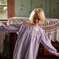 A young child wears the Louise Misha Baby Girls’ and Girls’ Anette Dress, a light purple corduroy style featuring an embroidered yoke and delicate ric rac details, while standing indoors near a dresser draped with lace. Painted flowers adorn the wall behind her.