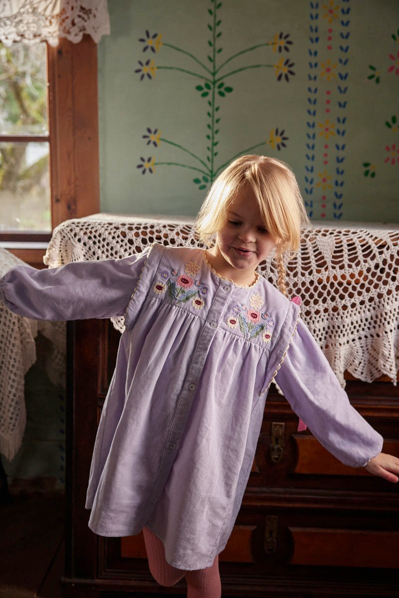 A young child wears the Louise Misha Baby Girls’ and Girls’ Anette Dress, a light purple corduroy style featuring an embroidered yoke and delicate ric rac details, while standing indoors near a dresser draped with lace. Painted flowers adorn the wall behind her.