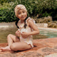 A child in a Louise Misha Andrea Bathing Suit and head wrap sits poolside, smiling with hands on lap, surrounded by lush greenery and the inviting pool.