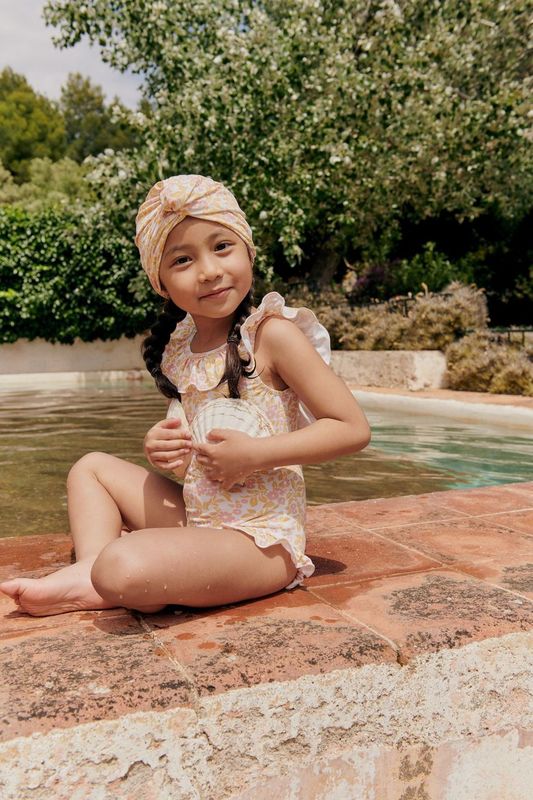 A child in a Louise Misha Andrea Bathing Suit and head wrap sits poolside, smiling with hands on lap, surrounded by lush greenery and the inviting pool.