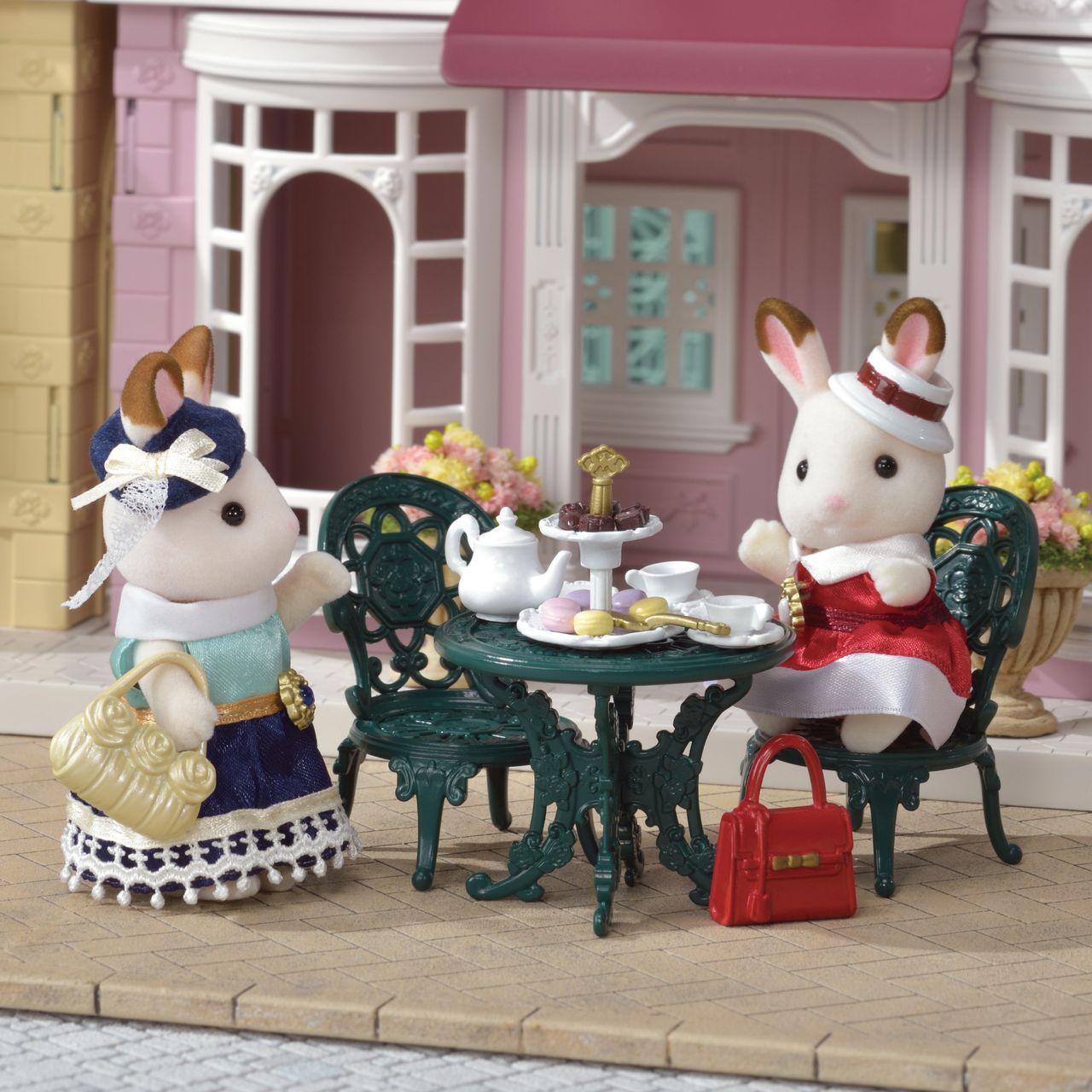 Two Calico Critters rabbits from the Tea and Treats Set enjoy tea, pastries, and handbags at a small outdoor café garden table surrounded by delightful tea party accessories, in front of a charming pink and white building.