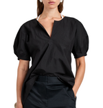 A woman stands confidently with one hand in her pocket, wearing the Proenza Schouler White Label Tia Top—a black, short-sleeve cotton-nylon blouse with a notched neckline—and black pants.