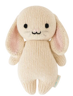 The Oatmeal Bunny - Baby 7" by Cuddle + Kind is a cream-colored, handcrafted knit plush bunny featuring long ears, black eyes, a stitched nose and mouth, and a small fabric tag on its side.