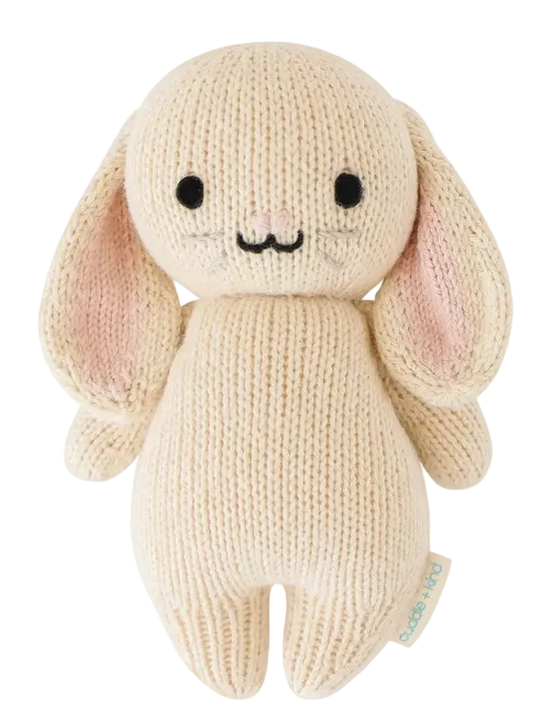 The Oatmeal Bunny - Baby 7" by Cuddle + Kind is a cream-colored, handcrafted knit plush bunny featuring long ears, black eyes, a stitched nose and mouth, and a small fabric tag on its side.