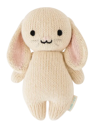 The Oatmeal Bunny - Baby 7" by Cuddle + Kind is a cream-colored, handcrafted knit plush bunny featuring long ears, black eyes, a stitched nose and mouth, and a small fabric tag on its side.