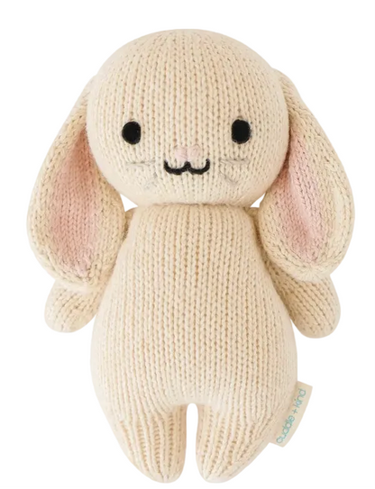 The Oatmeal Bunny - Baby 7" by Cuddle + Kind is a cream-colored, handcrafted knit plush bunny featuring long ears, black eyes, a stitched nose and mouth, and a small fabric tag on its side.