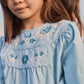 A young girl with straight brown hair wears the Louise Misha Girls’ Risa Blouse, a light blue top adorned with delicate floral embroidery and puffy sleeves, standing outdoors alongside another child.