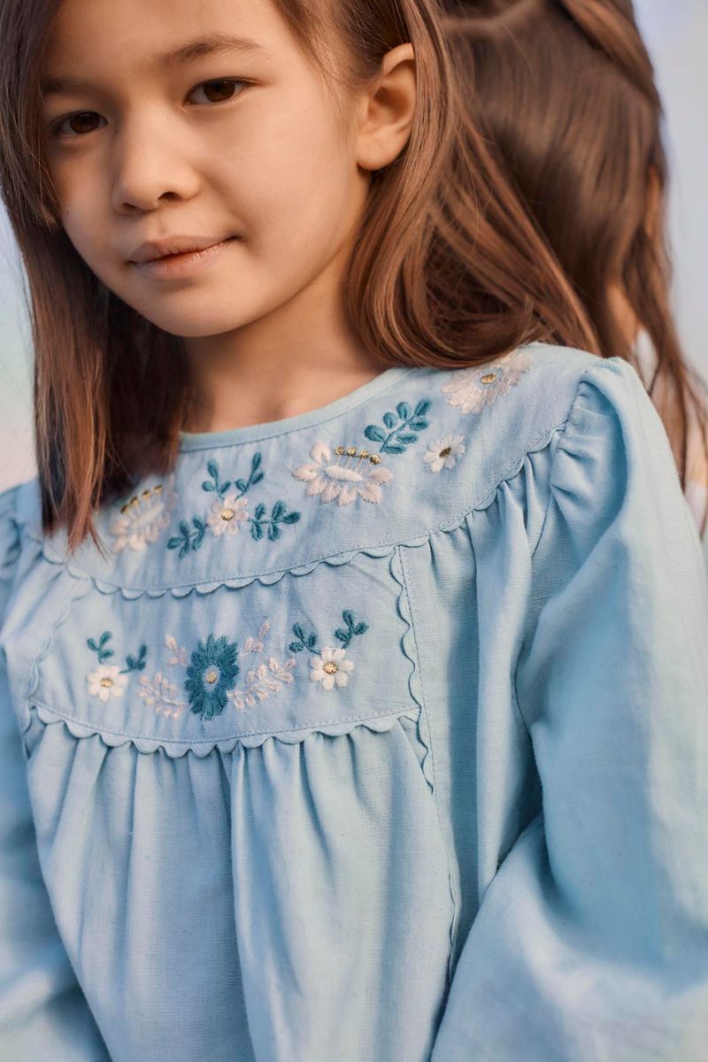 A young girl with straight brown hair wears the Louise Misha Girls’ Risa Blouse, a light blue top adorned with delicate floral embroidery and puffy sleeves, standing outdoors alongside another child.