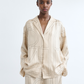 A person wears a loose-fitting beige Toteme Monogram Silk PJ Top with matching pants featuring a large abstract line pattern, standing against a plain white background.