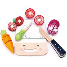The Mini Chef Chopping Board by Tender Leaf Toys features a smiling wooden chopping board, a wooden knife, and beautifully crafted toy vegetables—including a carrot, leek, onion, and tomato—all made from sustainable rubber wood.