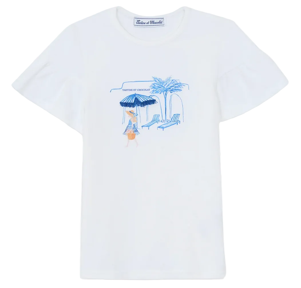 The Tartine et Chocolat Girls’ Beach Illustration T-Shirt is made from 100% cotton and showcases a delightful Santorini-inspired graphic in blue and orange, depicting a person lounging under a striped beach umbrella beside a chair, palm tree, and building. The design is completed with “Vanille et Chocolat” text above the illustration.