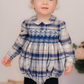 A toddler wears the Antoinette Boys' Louis Blue Tartan Romper with long sleeves and stands indoors, looking slightly upward.